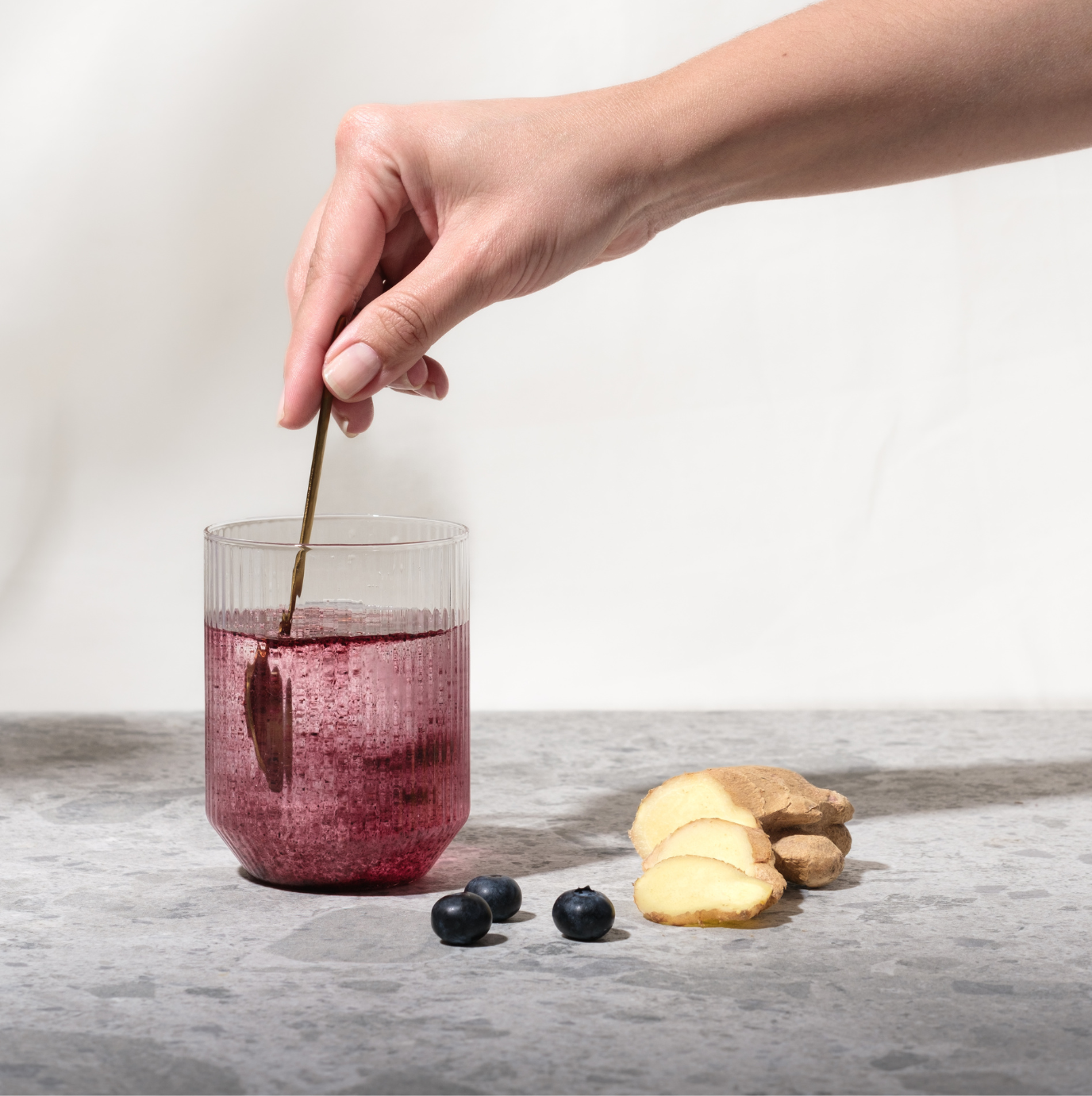 Ein Glas Wasser mit violettem myBioma Balance, das mit einem Teelöffel umgerührt wird – davor Heidelbeeren und Ingwer.